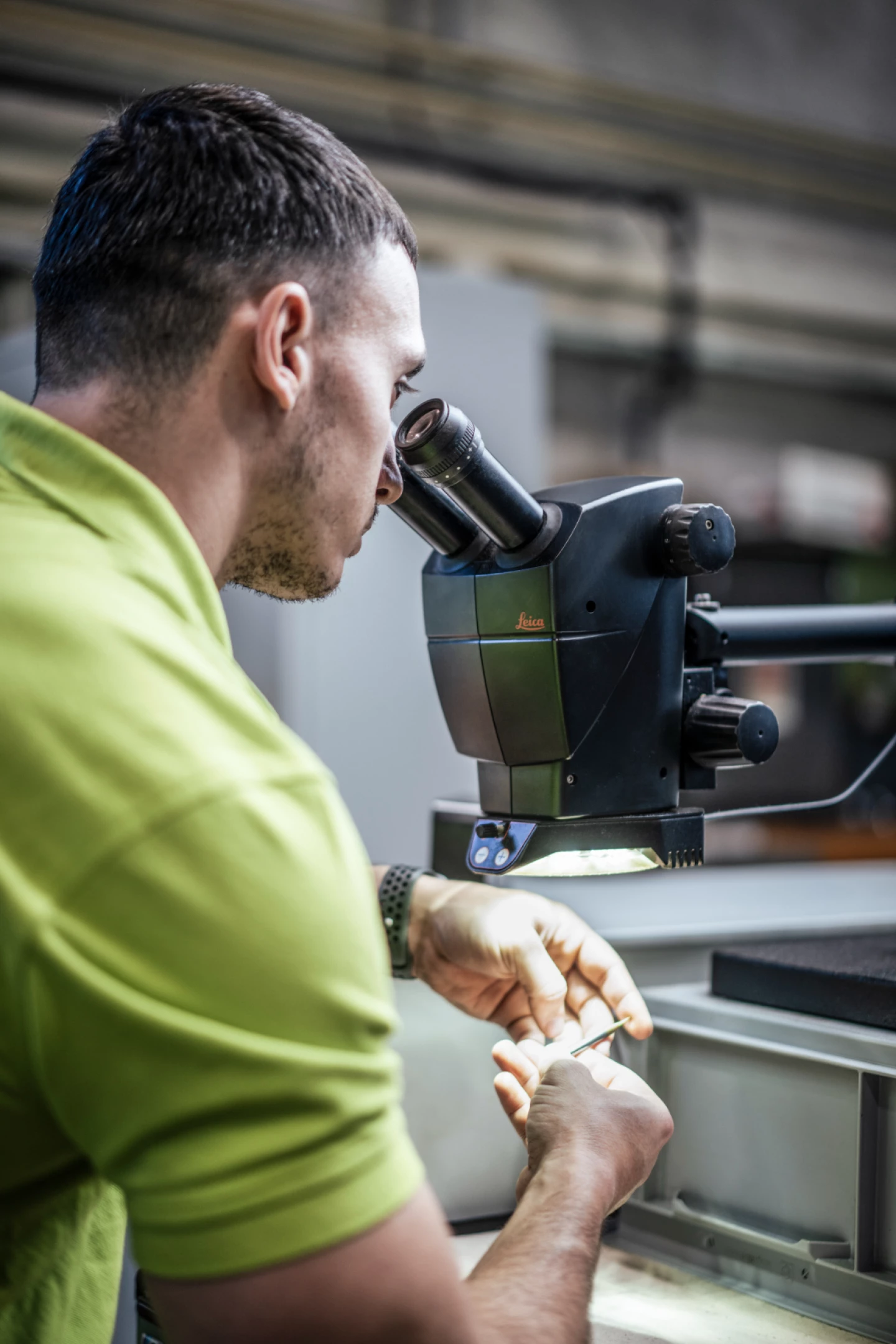 Ein Mitarbeiter begutachtet ein präzises Bauteil unter einem Mikroskop, um die Qualität zu prüfen. Die Kontrolle erfolgt mit einem Leica-Mikroskop, das von oben beleuchtet wird.