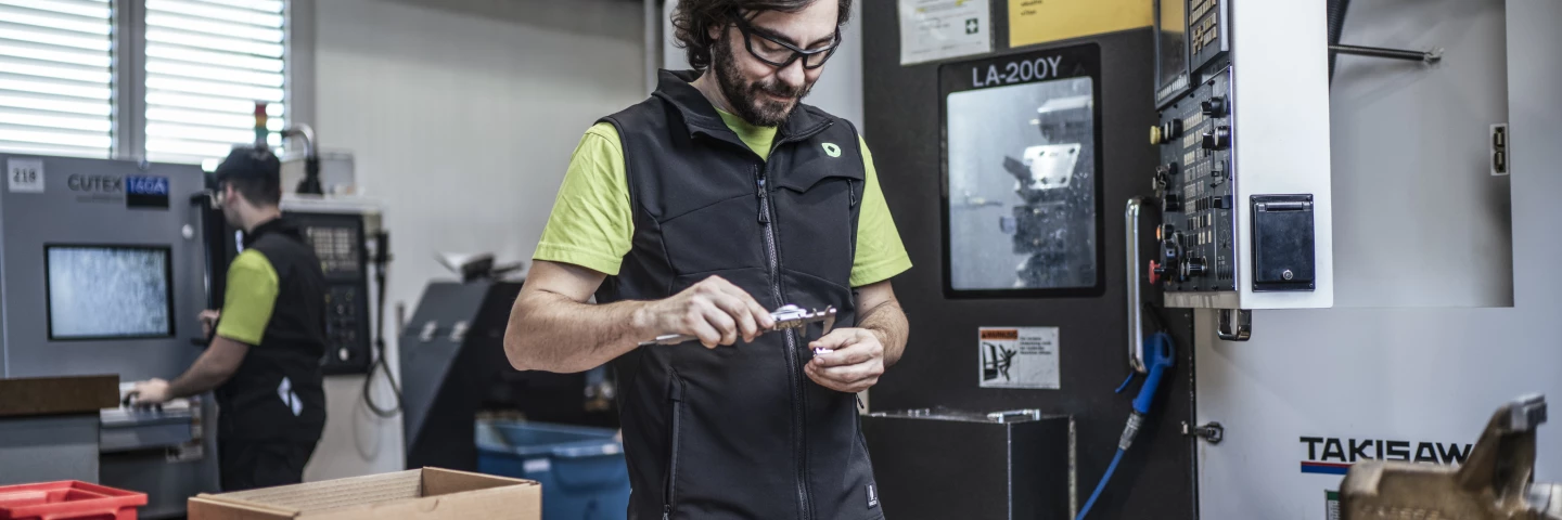 Person mit Schutzbrille und Arbeitsweste kontrolliert ein Bauteil an einer Werkbank; daneben liegen Werkzeuge und Kleinteile, dahinter sind CNC-Dreh-/Fräsmaschinen und weitere Beschäftigte in einer industriellen Fertigungsumgebung.